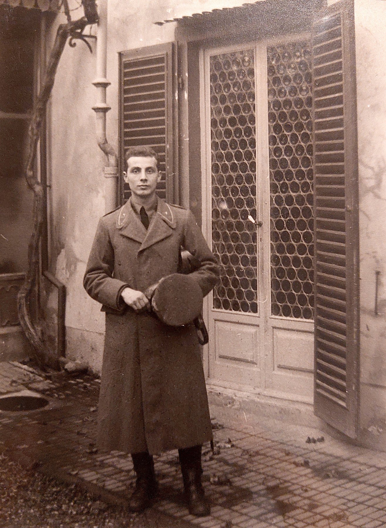 Aldo Grassi in cappotto militare davanti a una porta a vetri, 1939